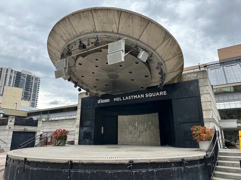 Mel Lastman Square's concrete bandshell stage under a cloudy sky, with residential towers and the North York Civic Centre roofline behind