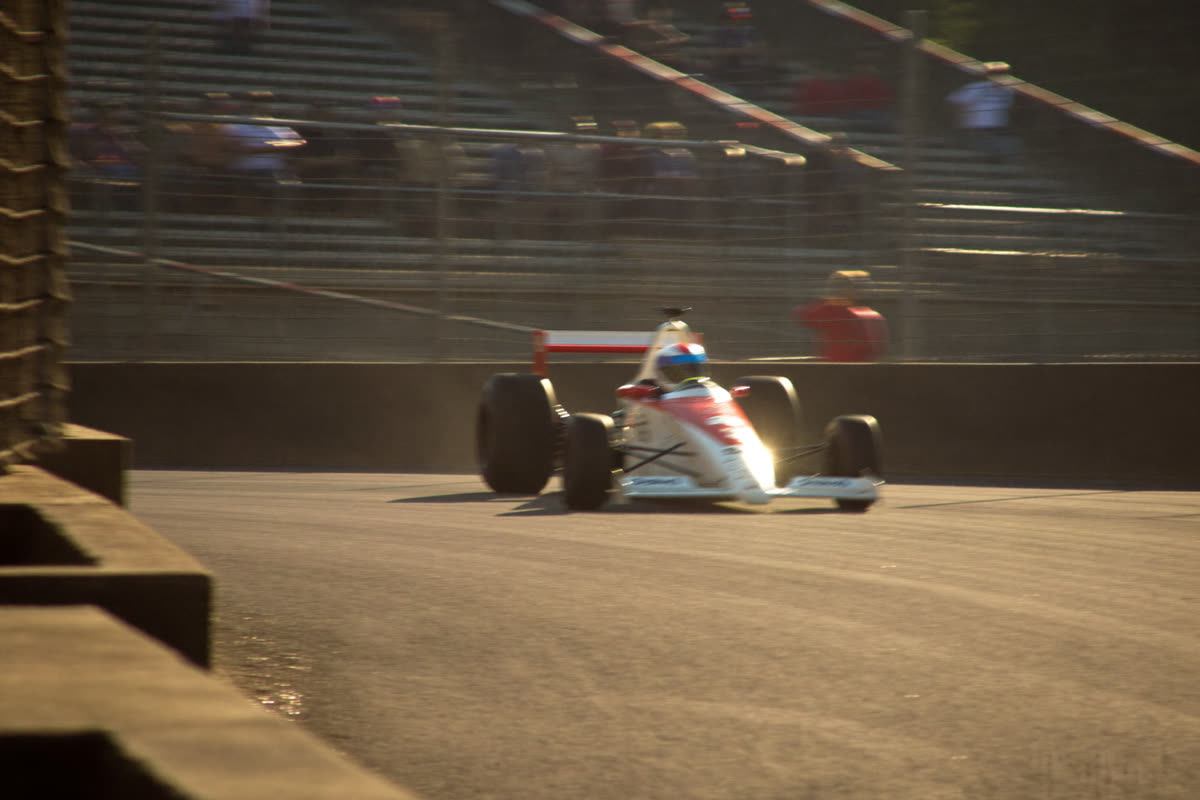An open-wheel race car on a temporary street circuit lined with concrete barriers and catch fencing, late-afternoon light